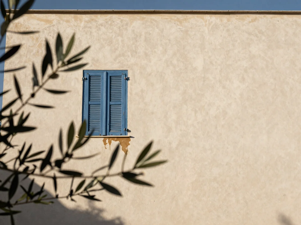 Candid close view of a pale rendered Mediterranean house wall in soft winter light, a small patch of damp shadow near a
